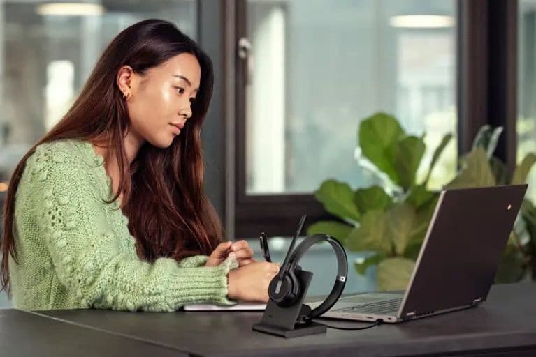 Someone working at a laptop, with the EPOS IMPACT 5000 Go headset on its charging stand beside it